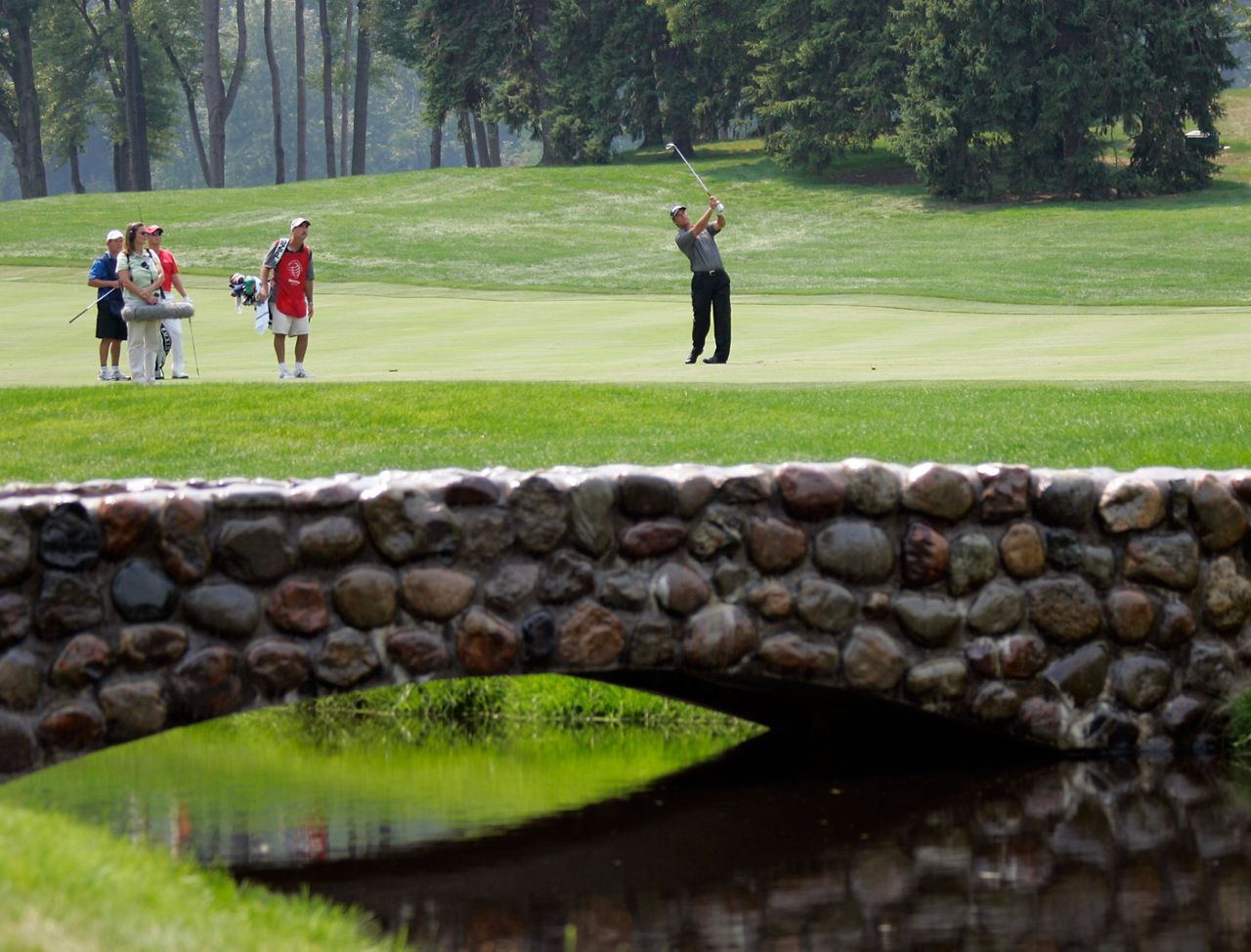 A golfer swings at a all beyond a stone bridge on a green course